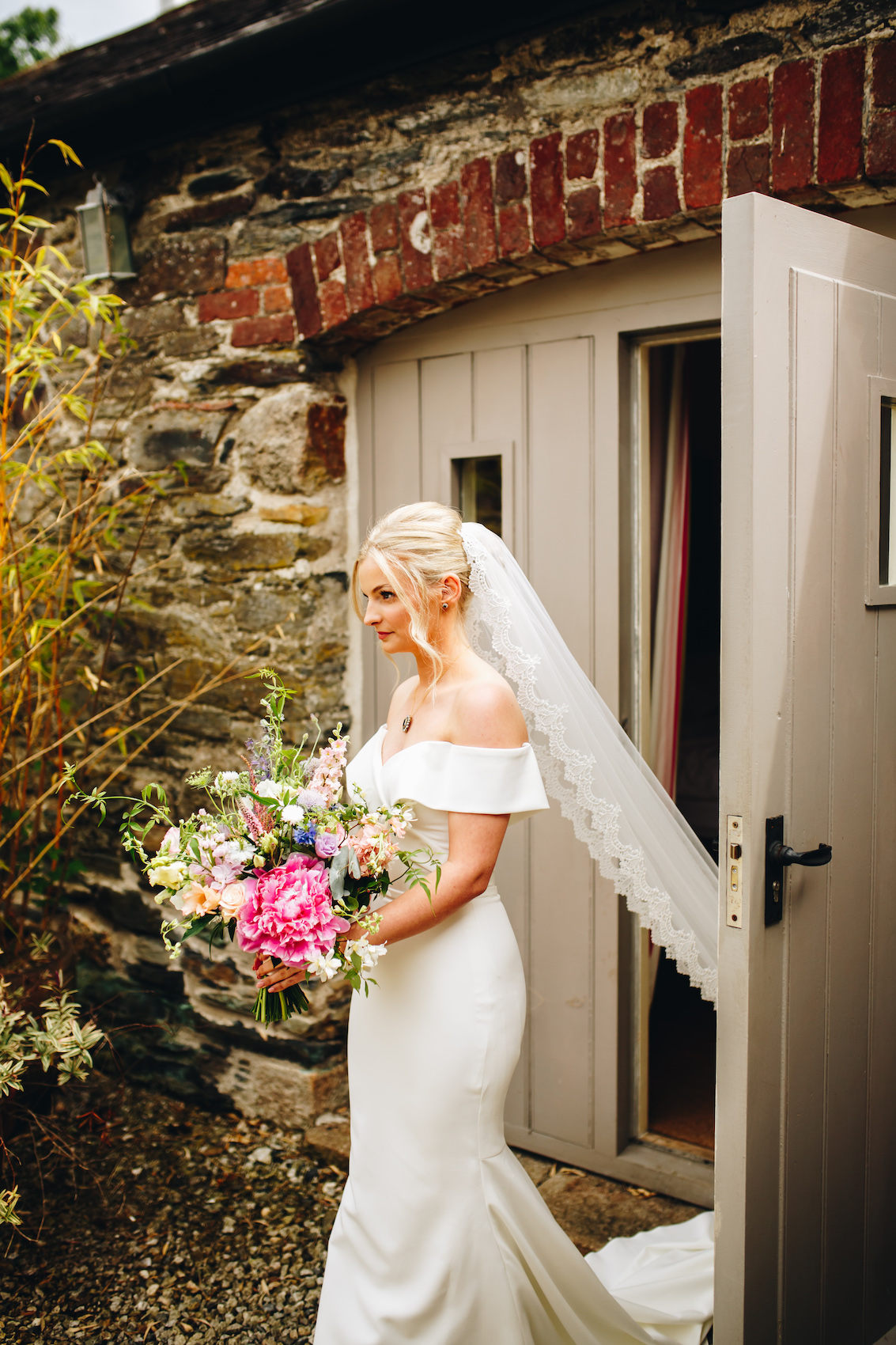 Bride exiting the bridal prep area to get married - wearing an off-the-shoulder dress with a sweetheart neckline, veill flowing behind her, holding a beautiful large bouquet