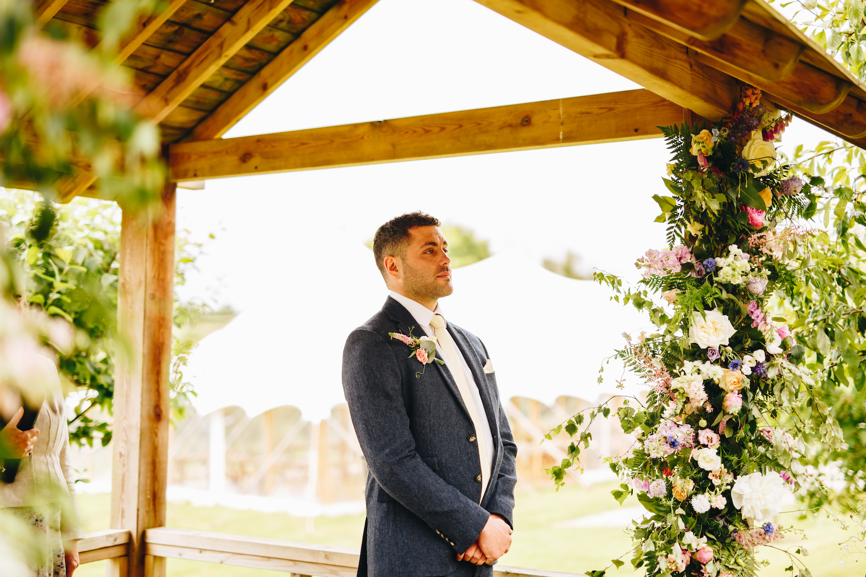 Groom tears up as he sees his fiancée in her wedding dress for the first time, in a wooden pergola adorned with florals