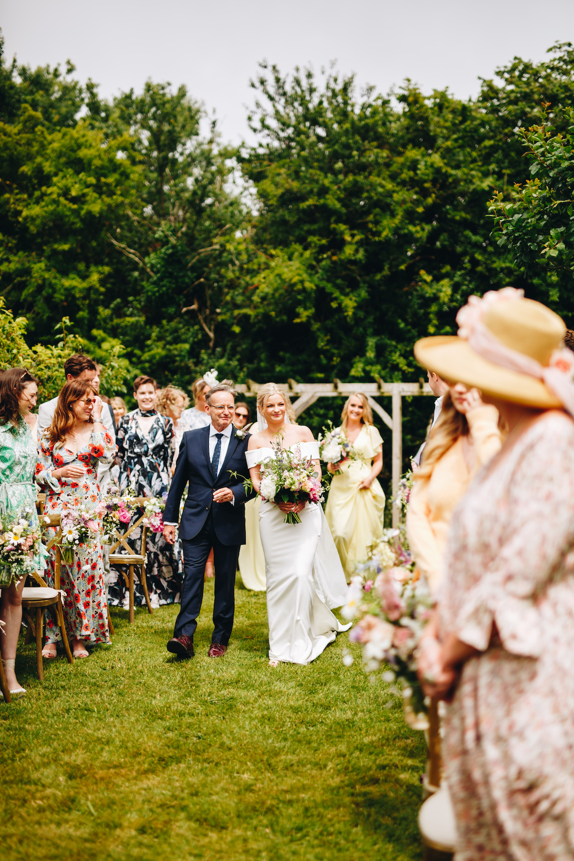 Bride and her dad walk up the aisle at an outdoor wedding with trees in the background