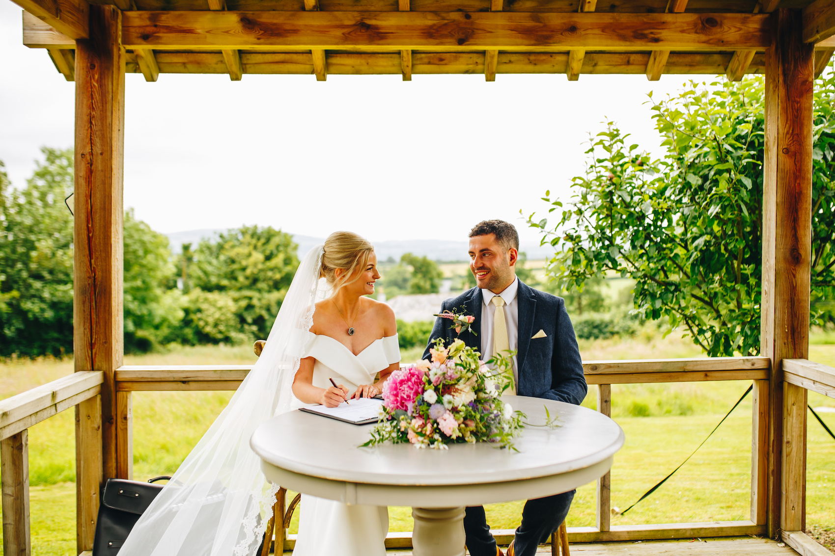 Bride and groom smile together after they've signed the register. Sat at a circular table outdoors under a wooden pergola