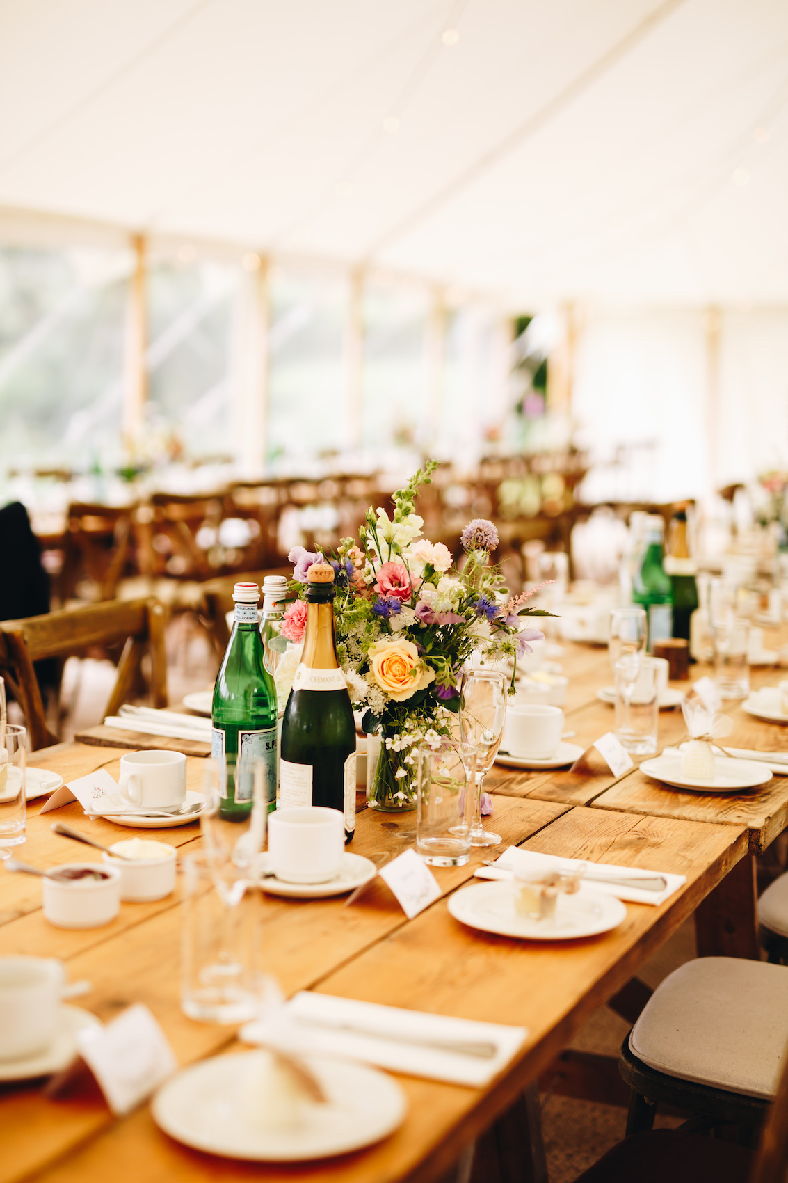 Close up of florals on long wooden banquet table, with tableware and fizz laid out too