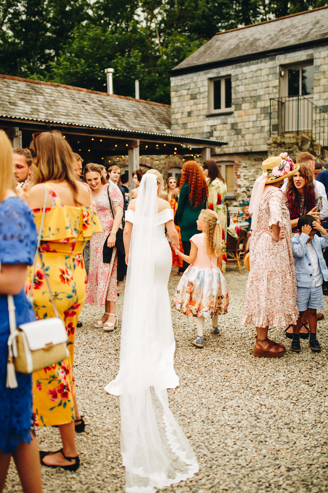 Bride walking away from the camera, in a gravel courtyard filled with guests in colourful outfits, as her veil flows behind her, holding the hand of a small girl who looks up at her