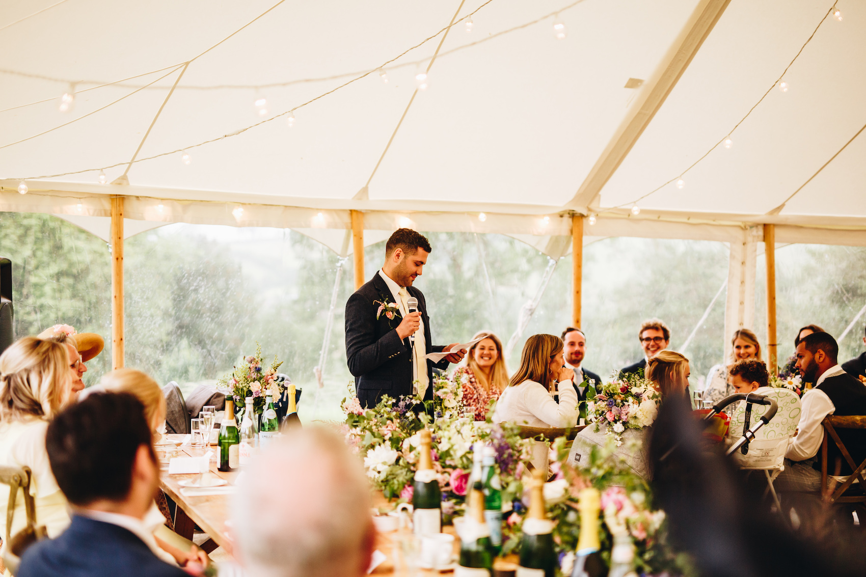 Groom gives a speech as his friends and family watch on smiling. The tables have floral displays and bottles of fizz and there are fairy lights in the top of the marquee