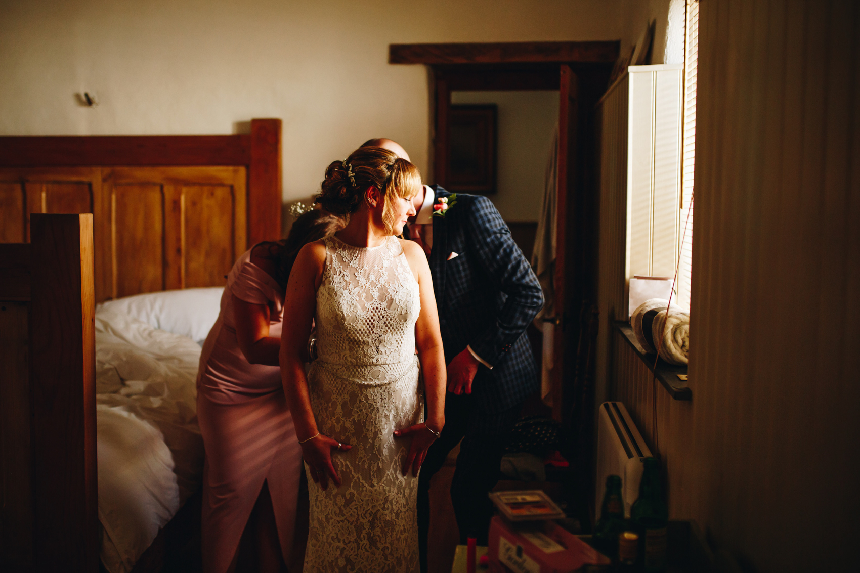 Bride's wedding dress being buttoned up by a bridesmaid and bridesman, looking over her shoulder towards the window