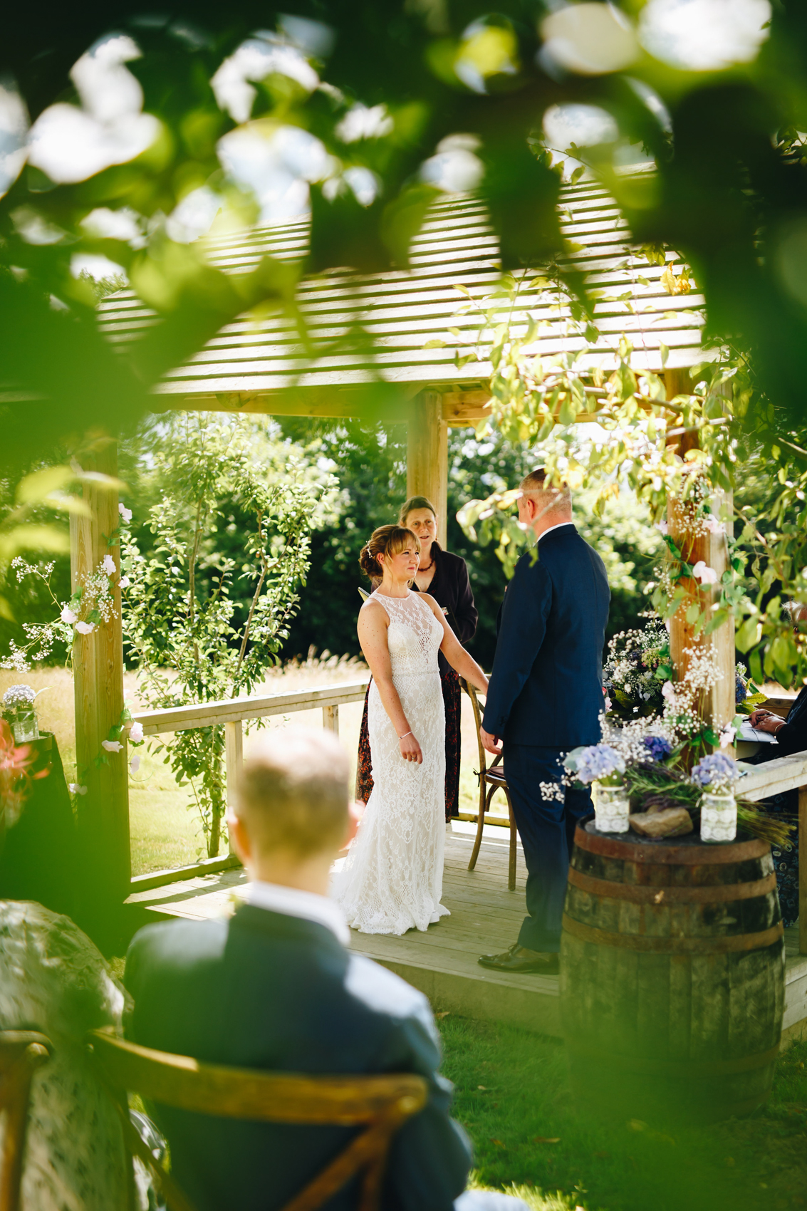 Bride smiling up at her groom, stood saying their vows, in a wooden pergola surrounded by trees and flowers