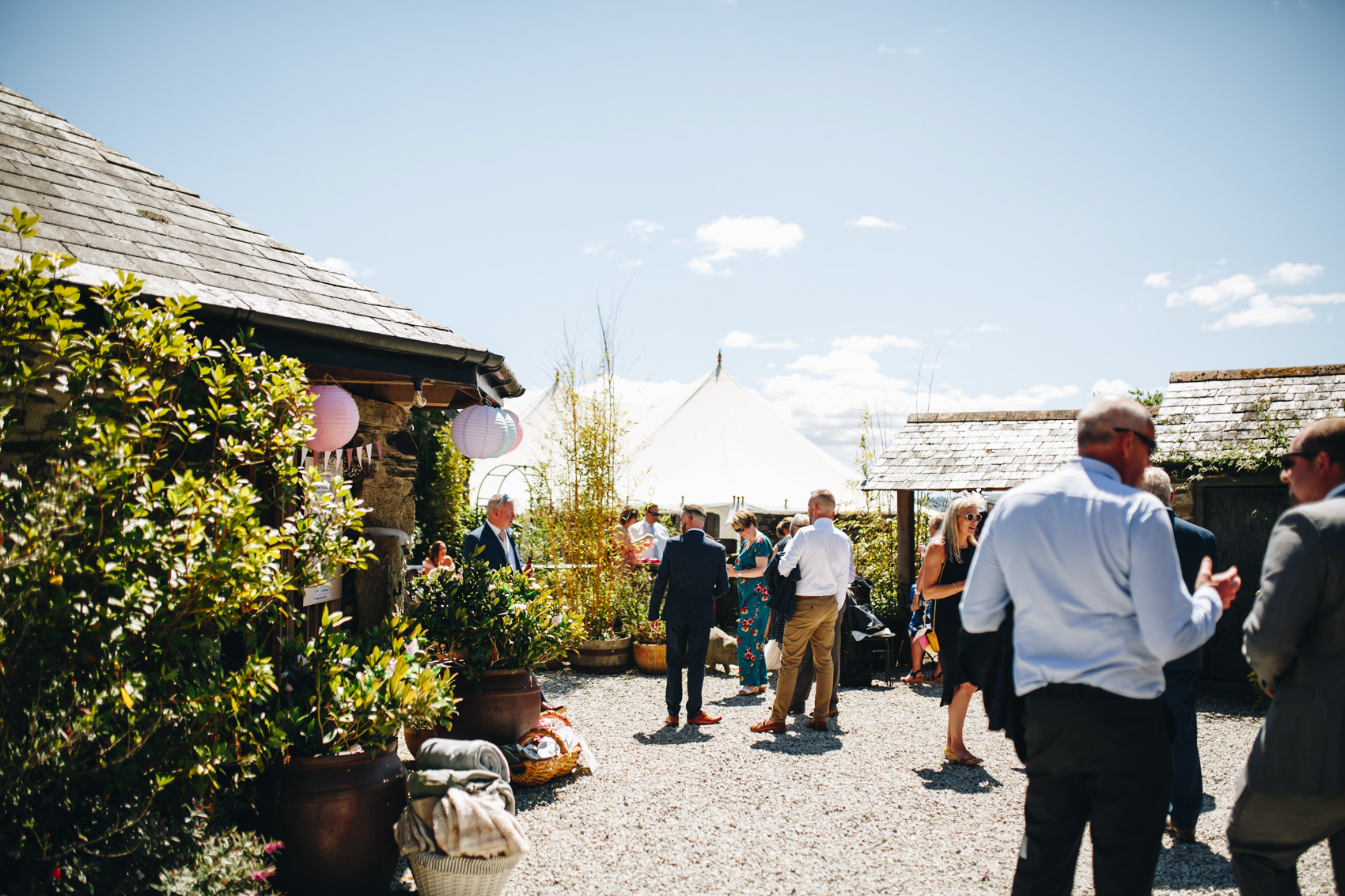 Guests mingling int he courtyard, with a marquee in the distance, and festoon lighting and potted plants all over