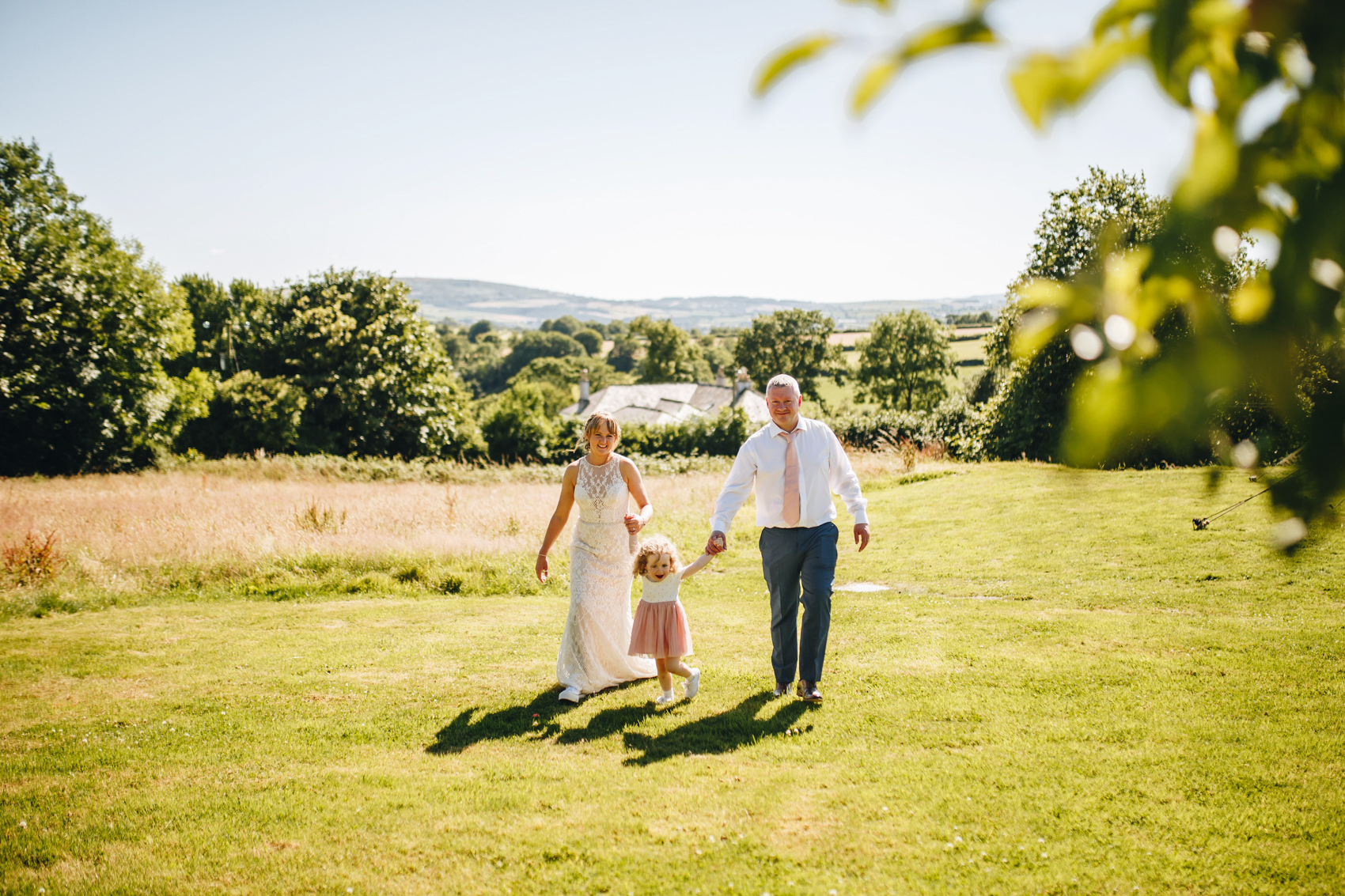Bride, groom, and their daughter walking hand in hand through the field towards the photographer