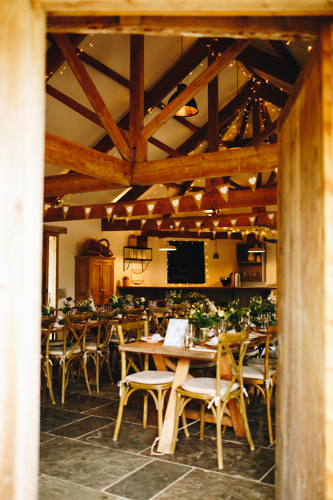 The view from a doorway of long wooden tables with chairs, with bunting and fairy lights in the beams, with greenery and flowers along the table amidst the plates and glasses