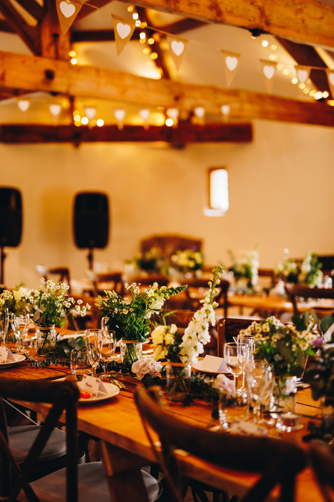 A close up of flowers on a wedding breakfast table - small glass vases with greenery, with fairylights on the beams above