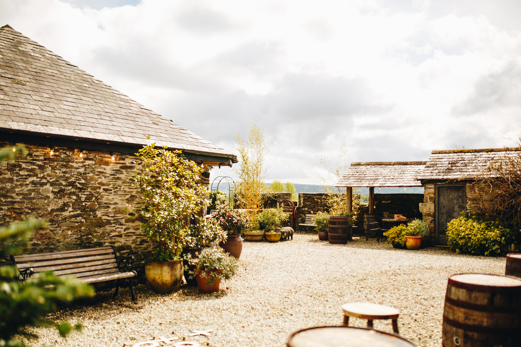 Wide photograph of a courtyard surrounded by low brick farmyard barn buildings and trees, with potted plants and seating dotted around