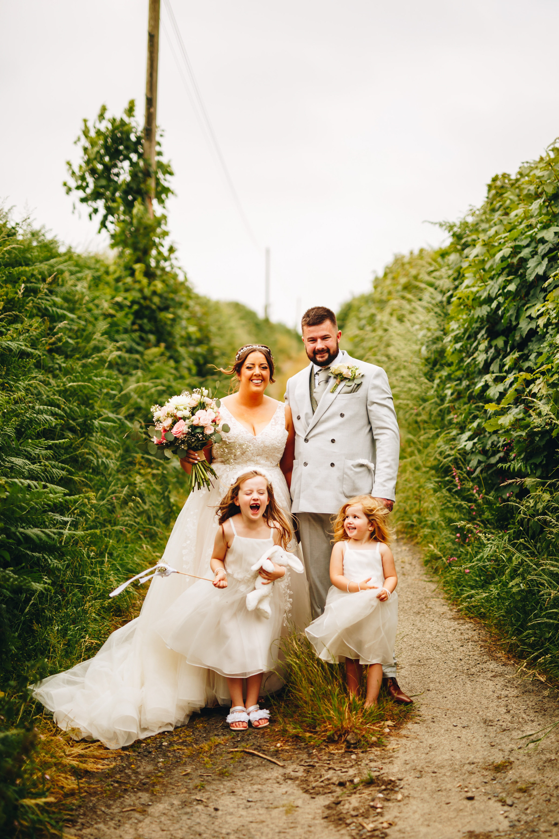 Bride, groom, and their two young daughters all gleefully happy, stood in a country lane in the breeze