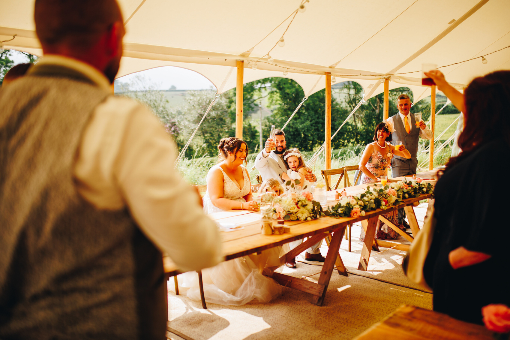 Bride and groom smile, groom holds their child and raises his drink, as the best man in the foreground gives his speech. Other guests raise their glasses too
