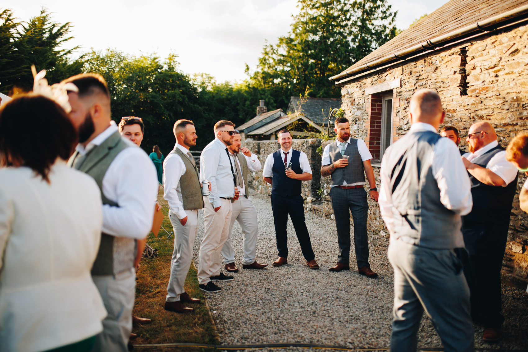 Groomsmen and guests stand around drinking and chatting in the evening sunlight in a courtyard
