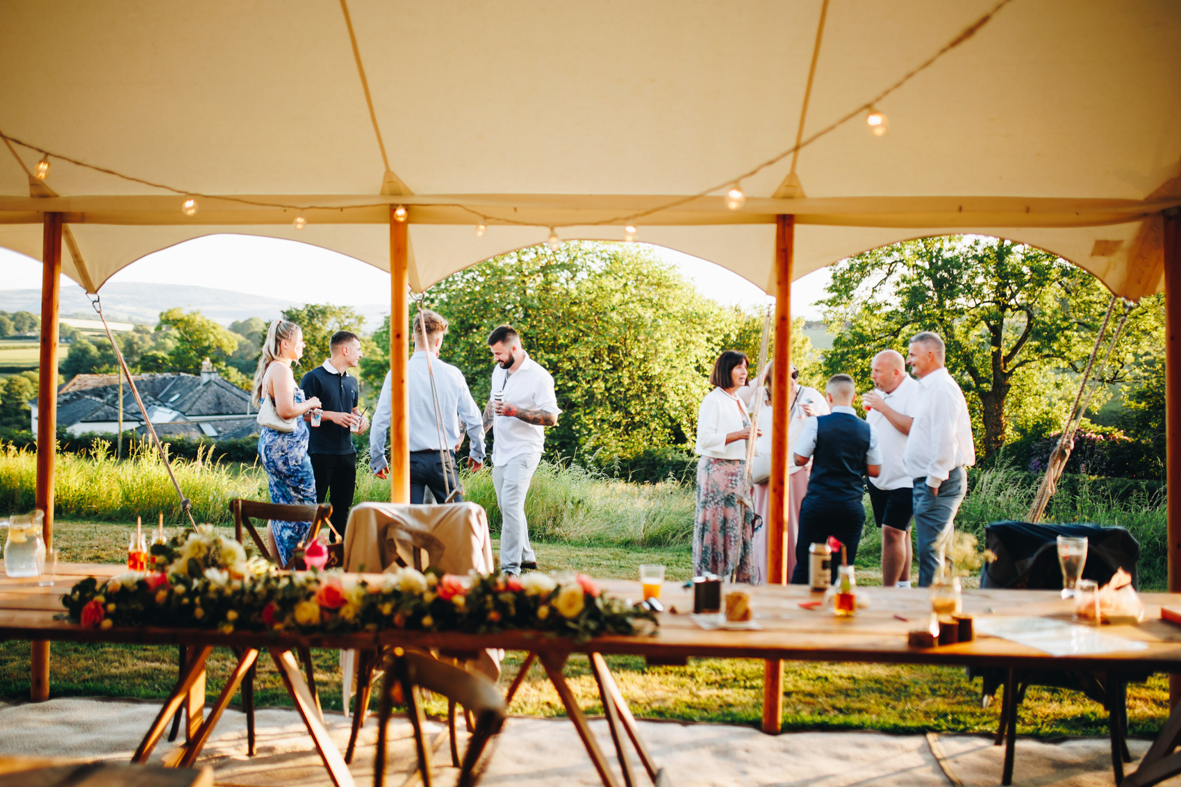 Sunny marquee with its sides open, guests mingling and drinking outside on the grass in the evening sunshine