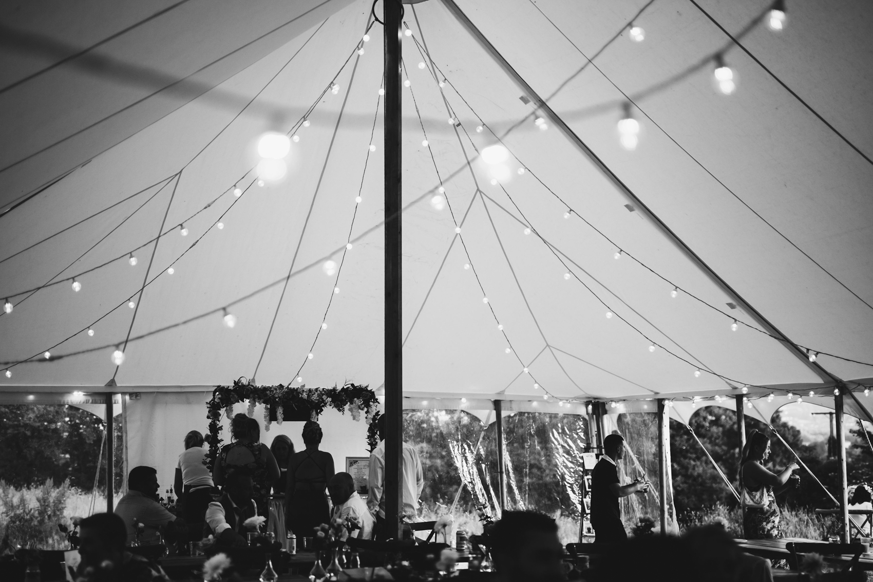 Black and white dark photo of marquee from inside, with fairy lights criss-crossing along its ceiling, with guests at the bottom of the frame at the bar