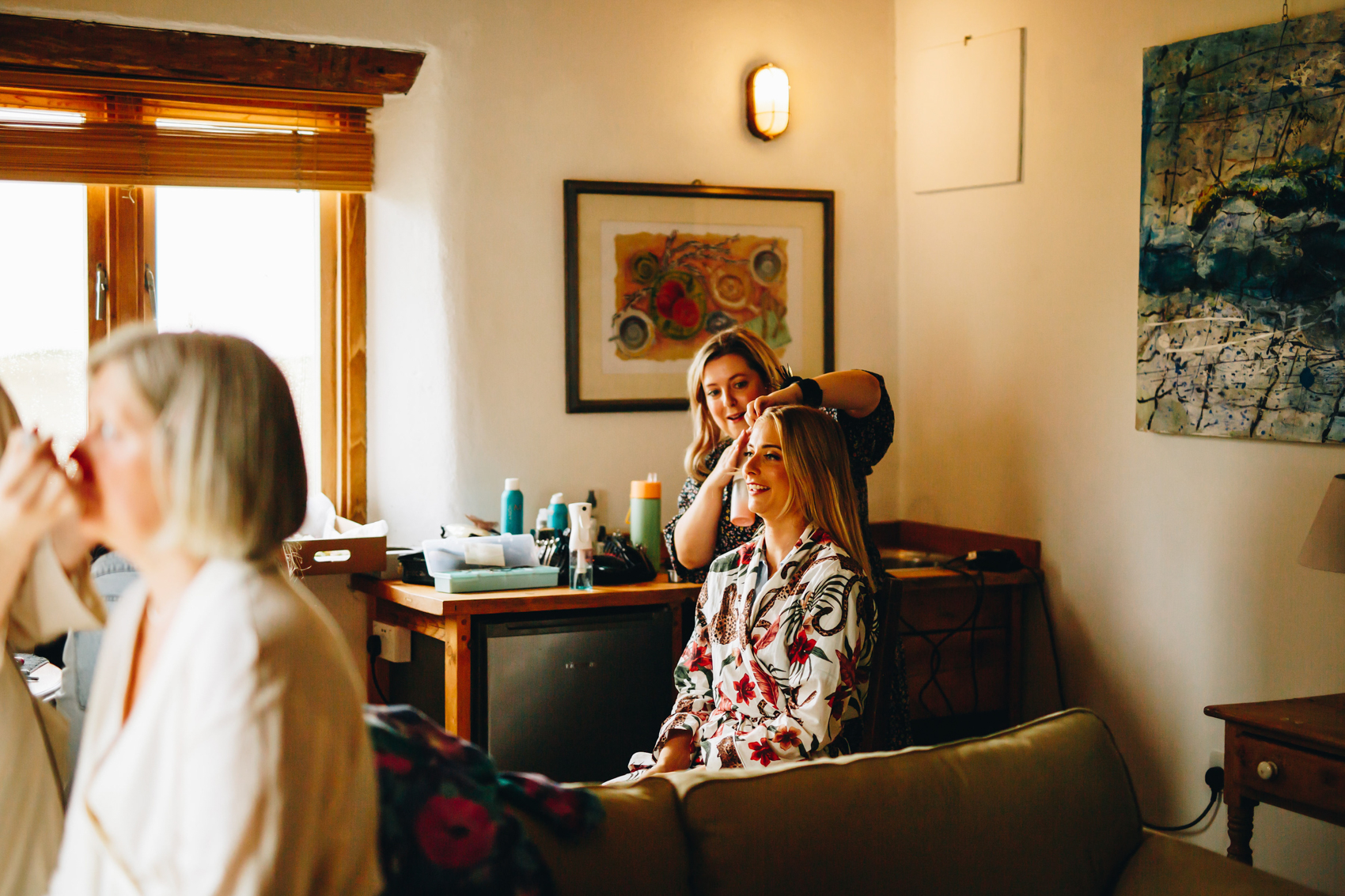 Photograph from across the room of bride having her hair done, with mum in the foreground having her makeup done
