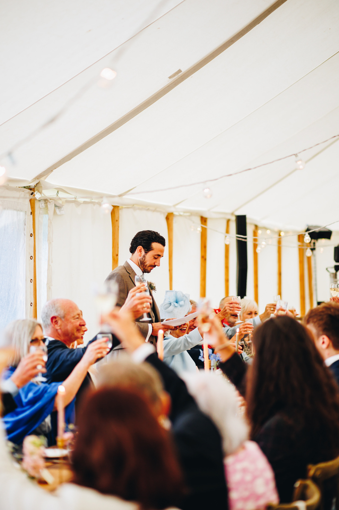 Groom stands amongst his seated guests who raise glasses at the speech he's making