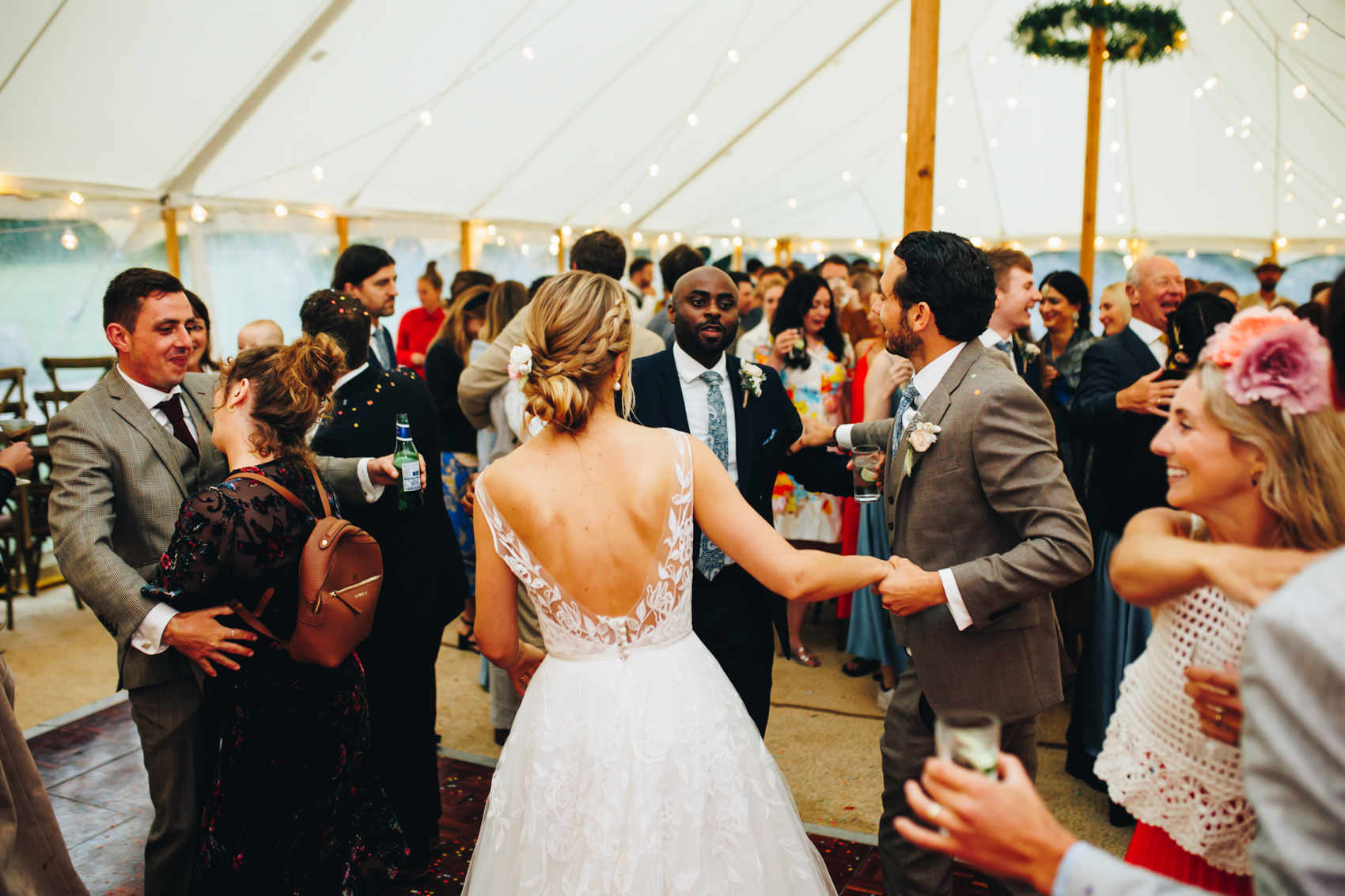 Bride and groom surrounded by their friends and family on the dancefloor at their wedding reception, with fairy lights in the background