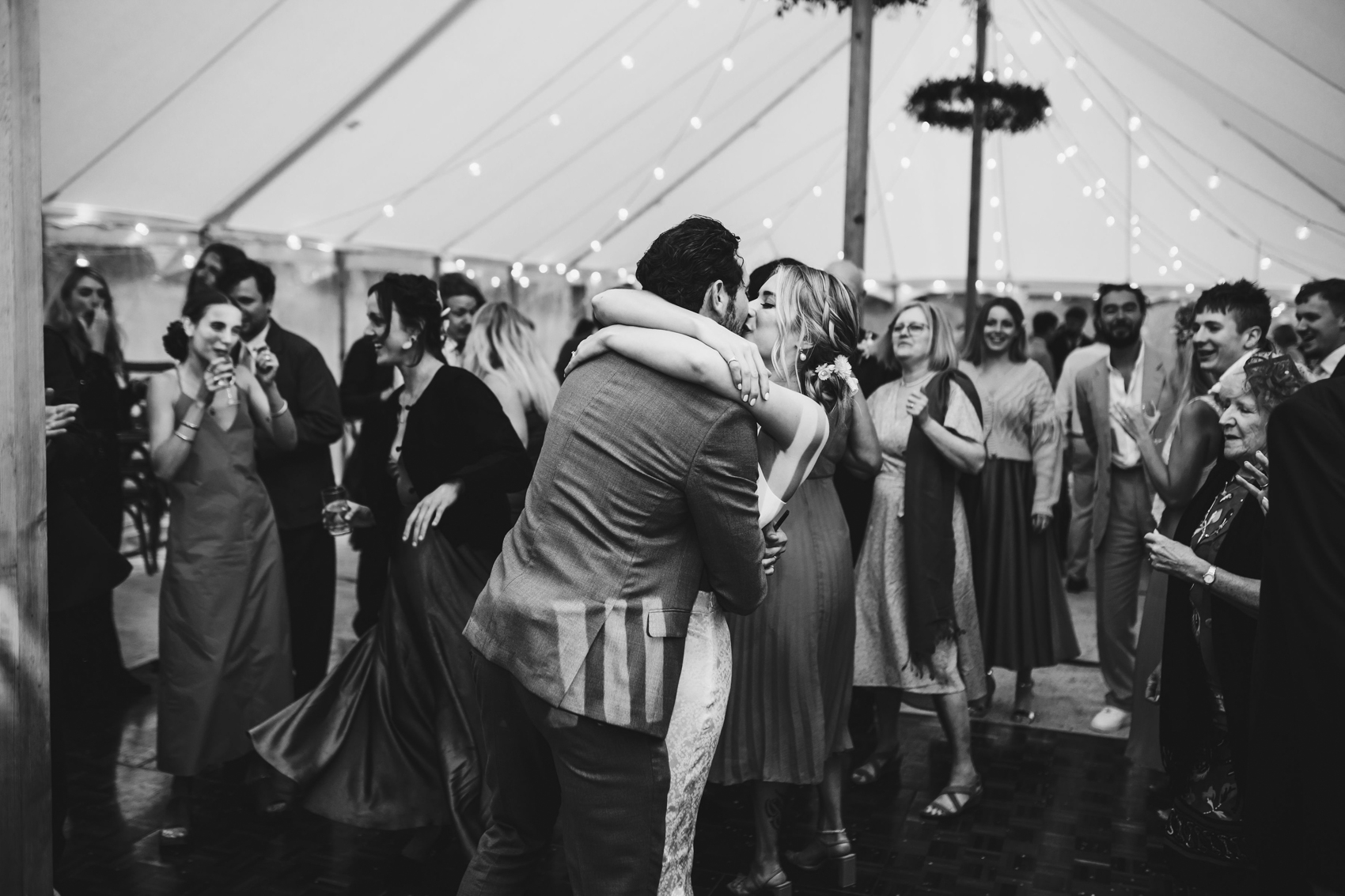 Black and white photo of wedding couple kissing on the dancefloor as their loved ones dance around them