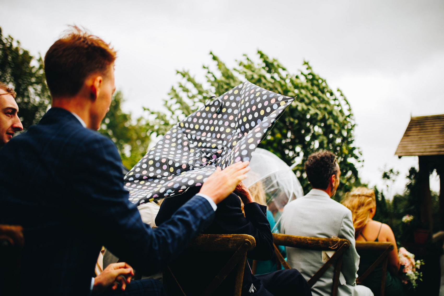 A guests holds an umbrella over their head that has blown inside out, while the man sat behind her holds its edge to stop it flying away. Trees in the background are being blown around too