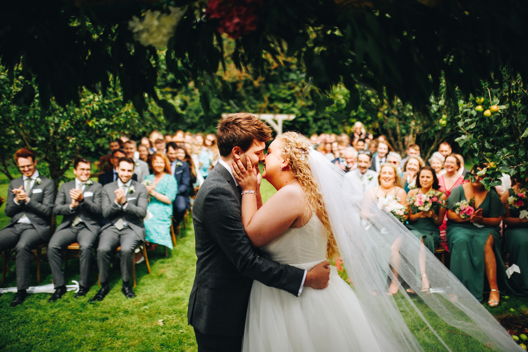Bride and groom's first kiss in front of their friends and family, under some greenery