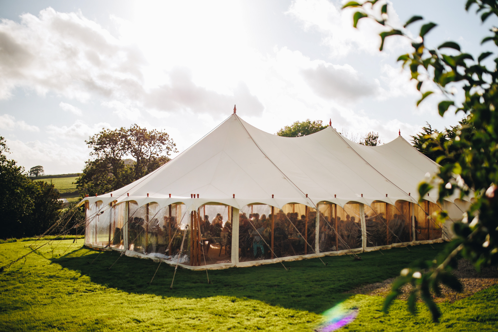 Photo of a marquee in a grassy field with trees in the foreground and blue cloudy skies above.