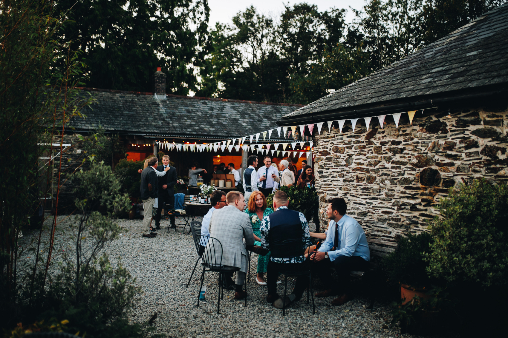 Wedding guests sit in the dim evening light with bunting attached to the brick barns surrounding them
