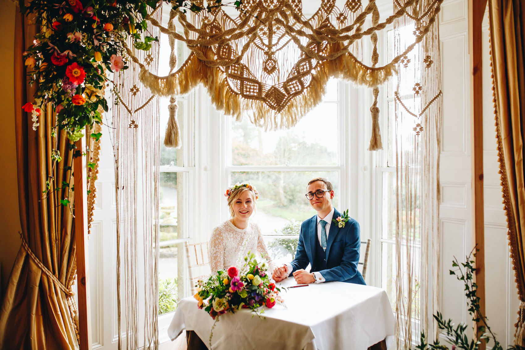 Bride and groom after signing the register and becoming legally married, sat at a table by a window and under their handmade macramé arch.