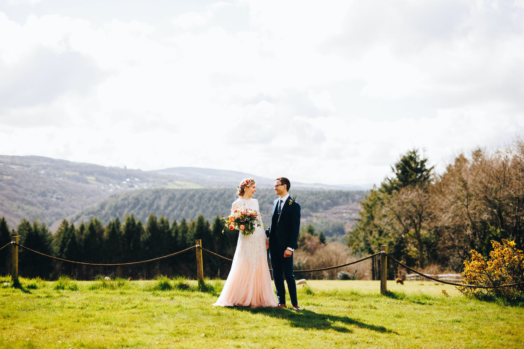 Bride, in pale pink dress with a flower crown and beautifully big bouquet, and groom in a blue suit with a button hole, stand with the valley in the background, smiling at each other and holding hands