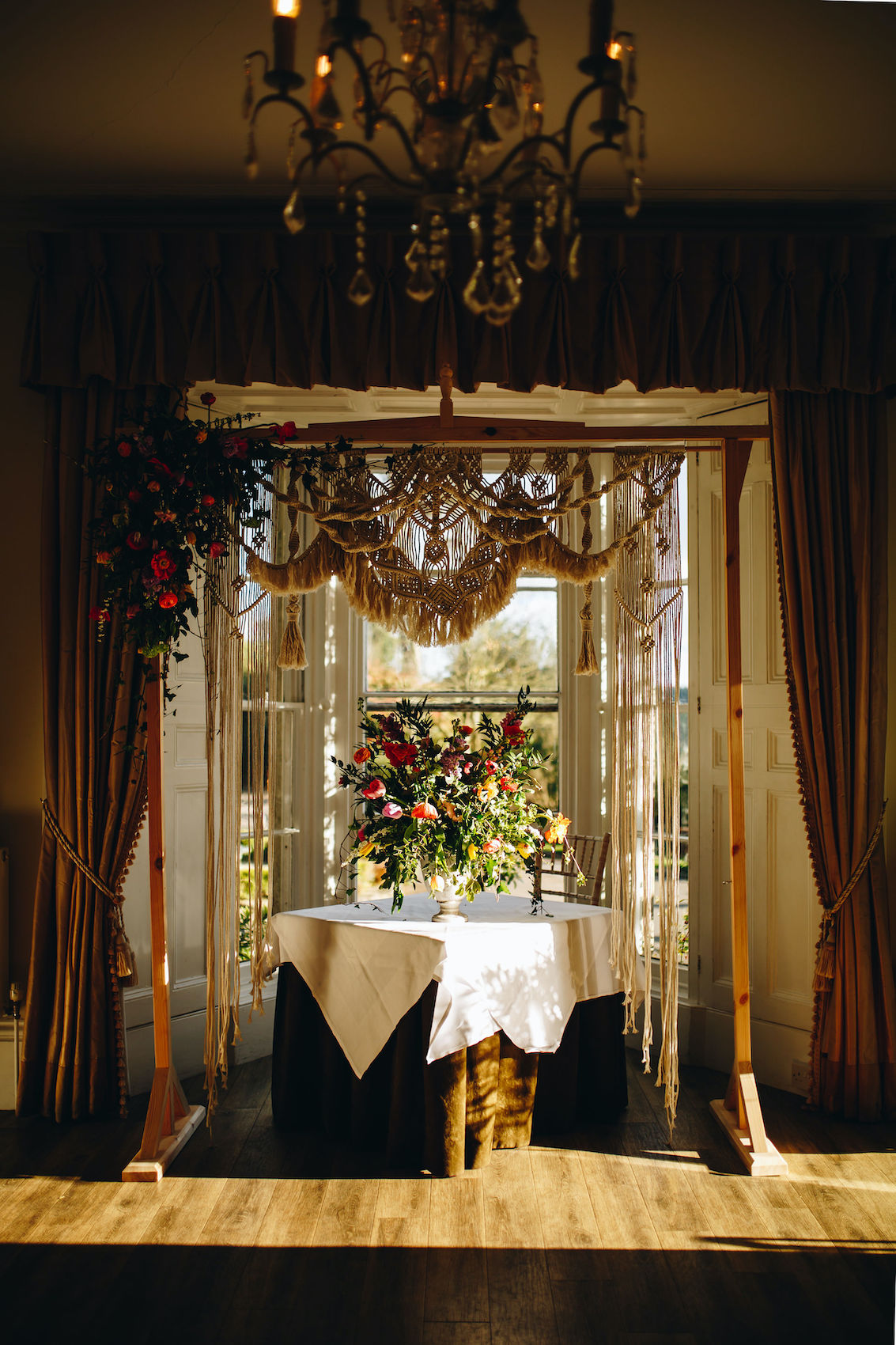 Photo of table under a macrame arch and in front of a window, with a massive floral display on the table, in the slanting evening sun coming in from the right side