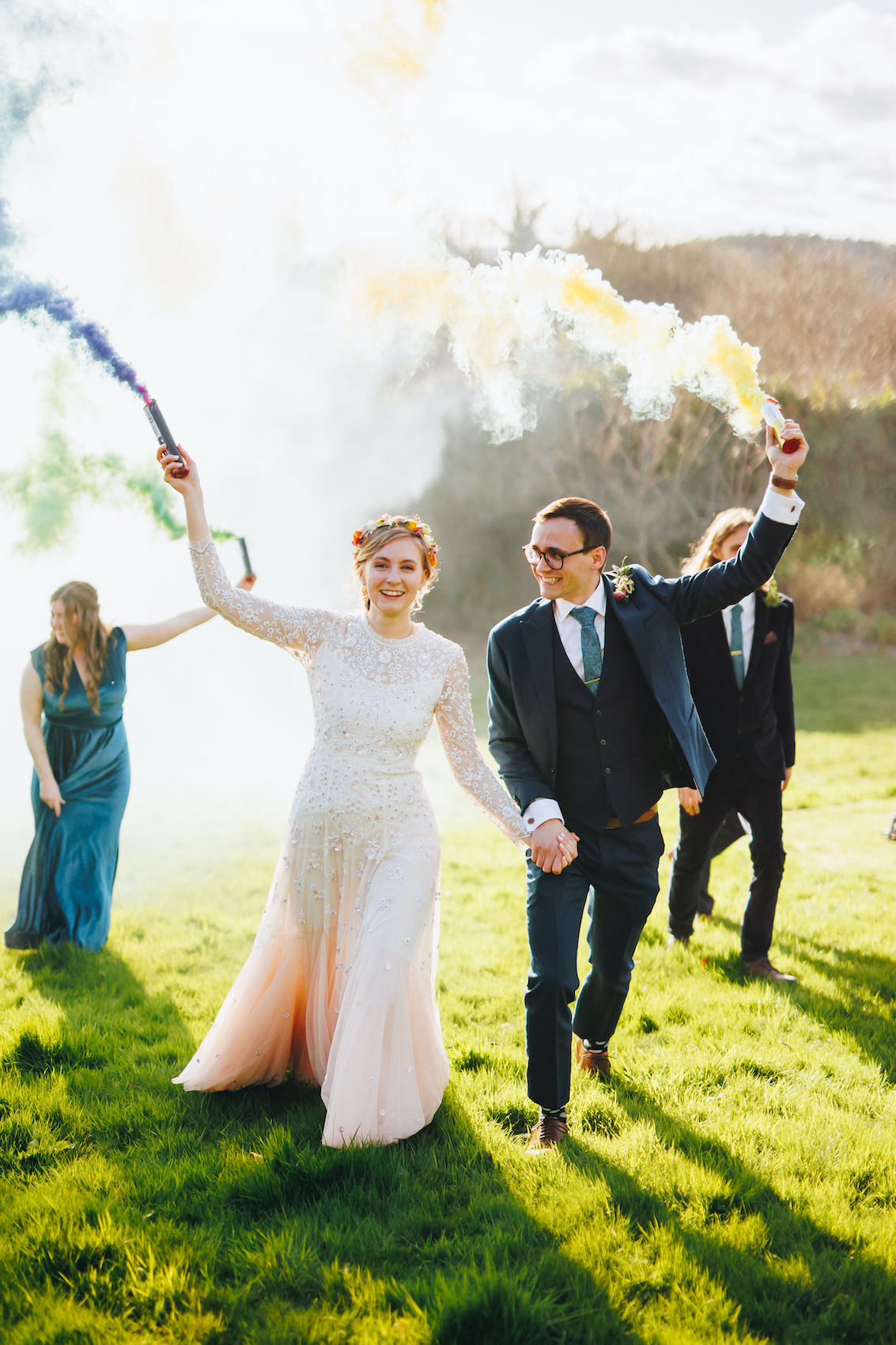 Bride and groom holding smoke bombs aloft in the evening light, with other members of the bridal party doing the same behind them