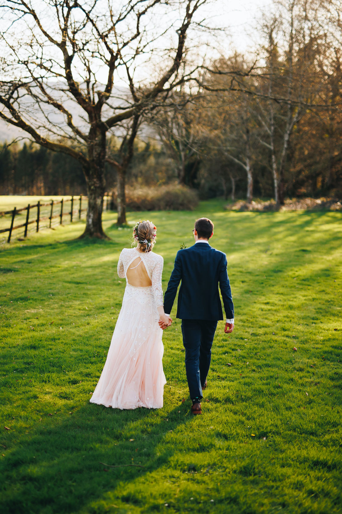 Bride and groom walk across a shadowy field towards the woodland