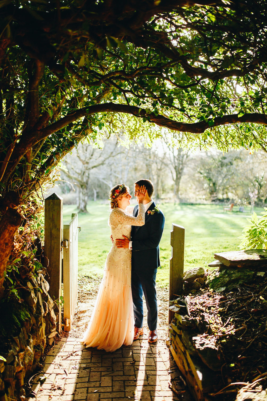 Bride and groom stand in a gateway, under a big old tree, backlit by the evening light, happily holding each other