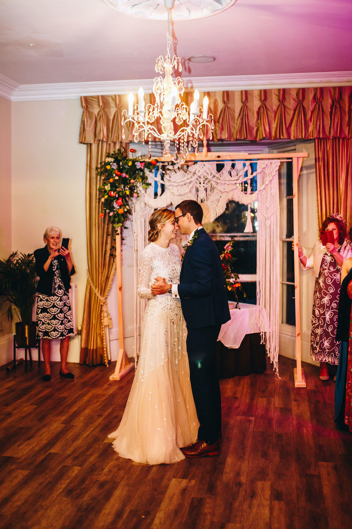 Newlyweds have their first dance, under a chandelier on wooden floor, smiling and holding each other close