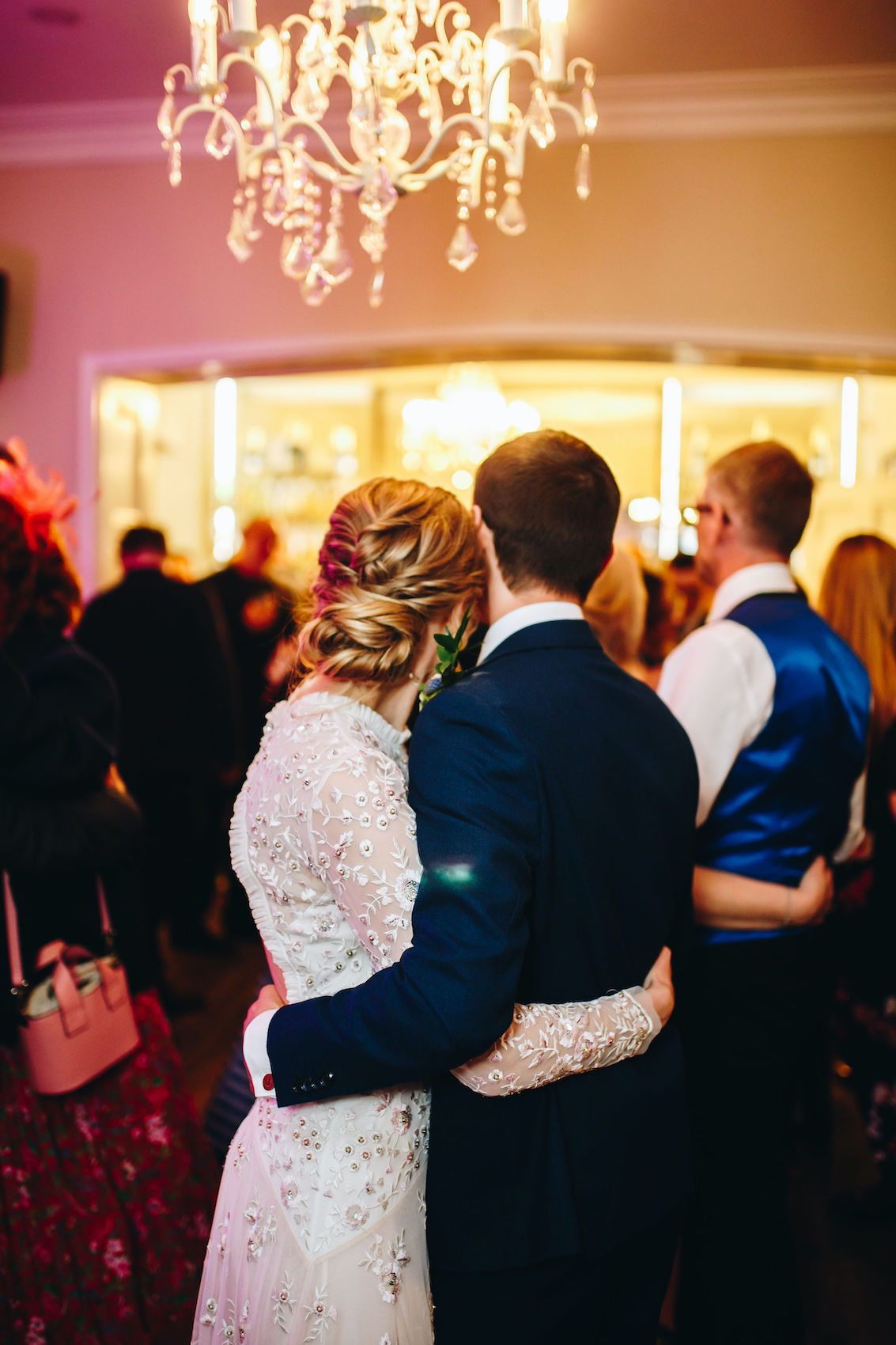 Newlyweds photographed from behind, arms around each other, looking out at their guests who have joined them on the dancefloor