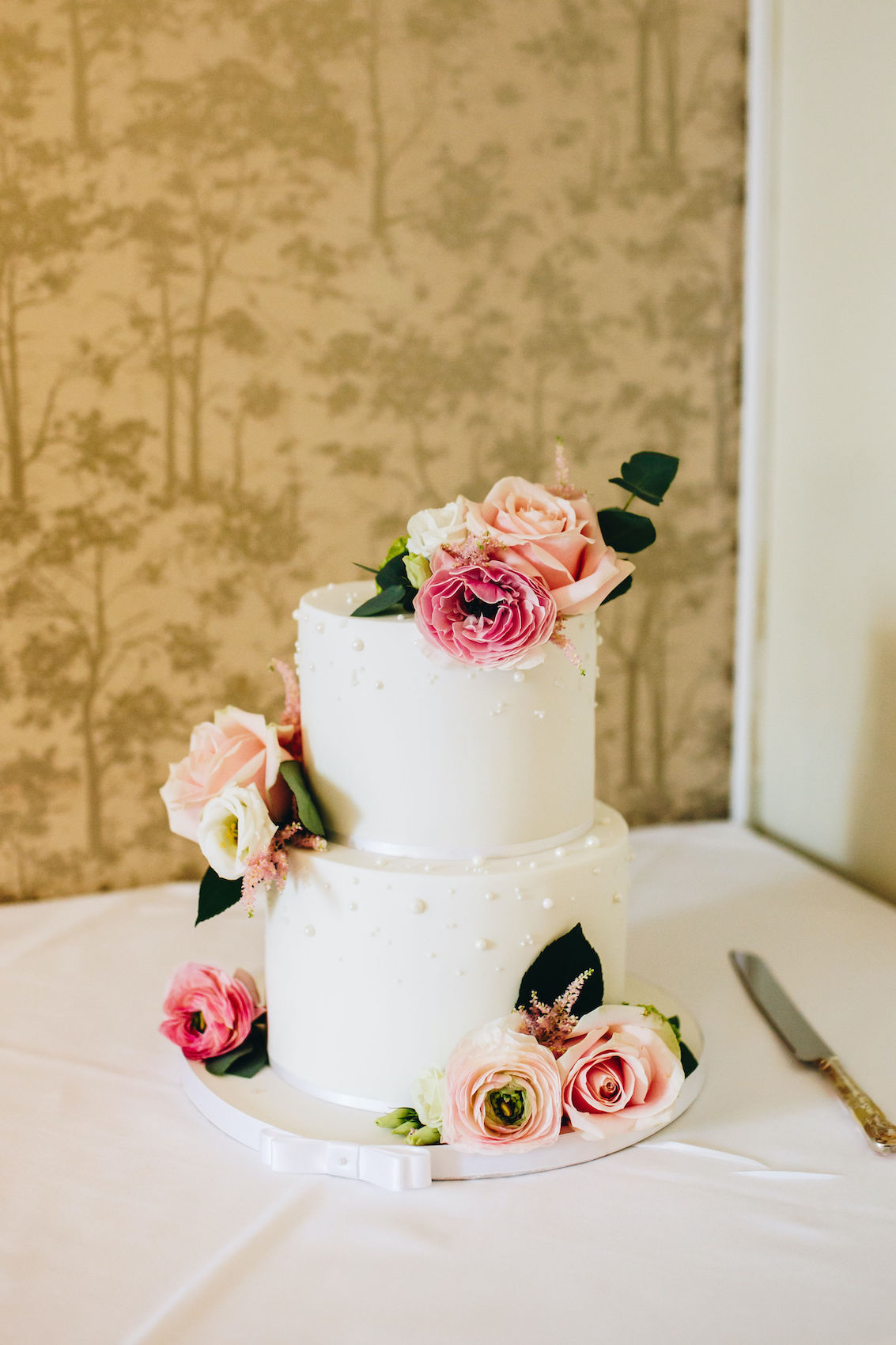 Photo of wedding cake, with white icing and pink flowers, sat on a table