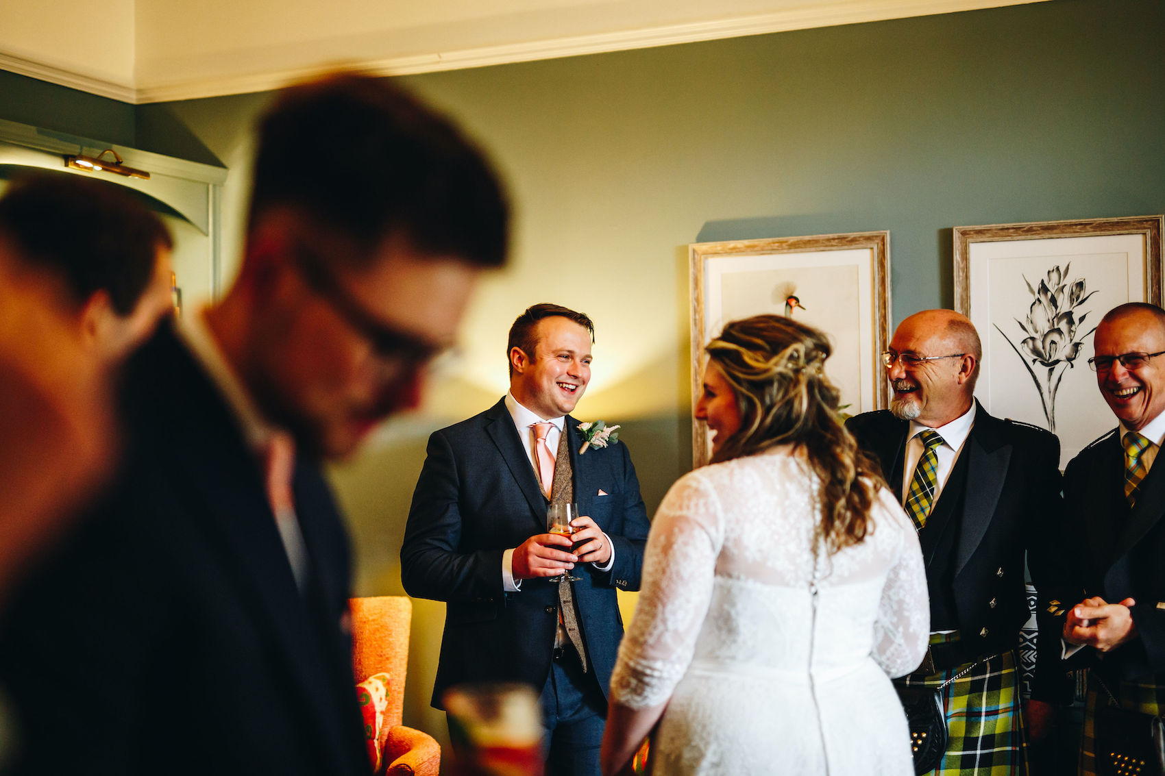 Groom laughs at something a guest has said, in a richly lit room with green walls and framed art