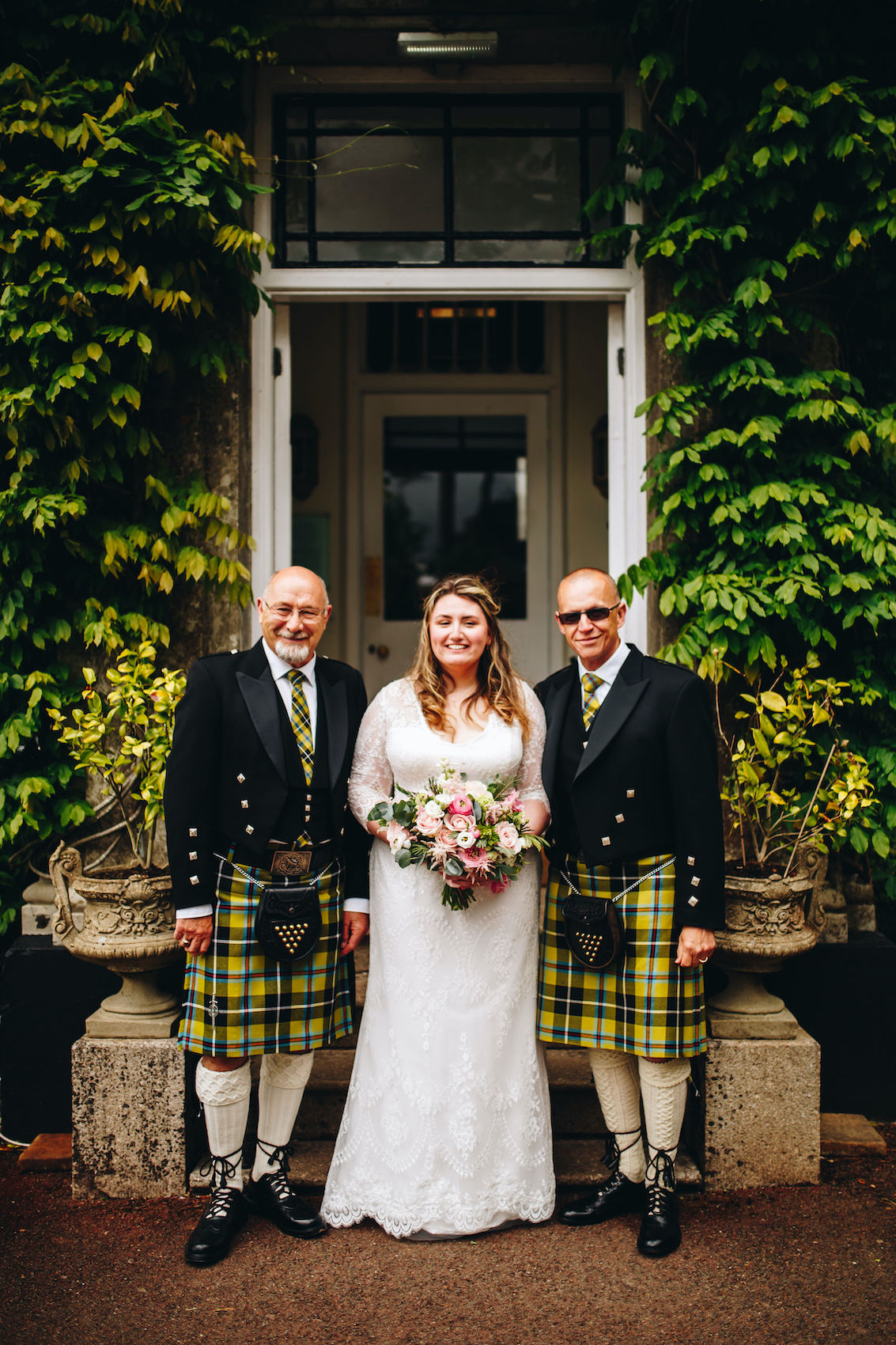 Bride stands with uncles who wear kilts, by the main door of a Manor Home adorned with wisteria leaves