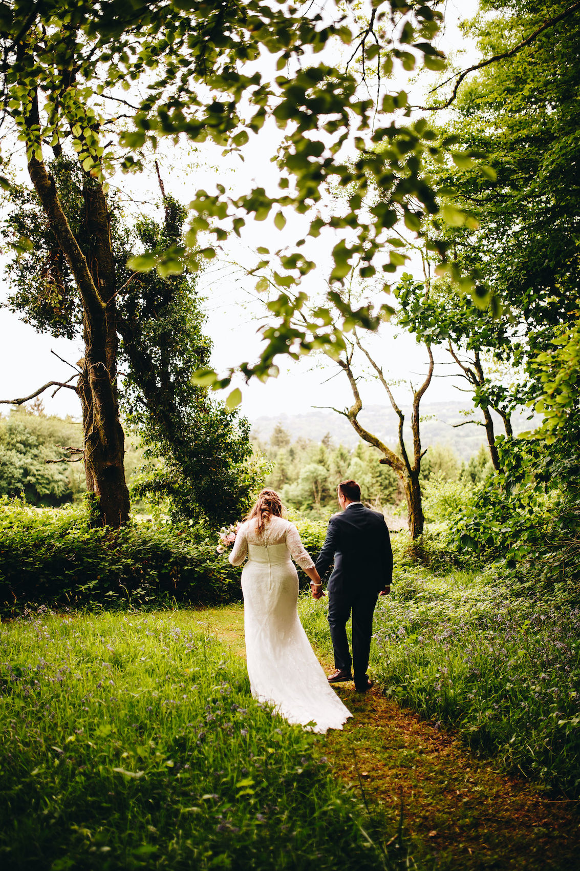 Bride and groom walking away from the camera hand in hand along a path through leafy trees