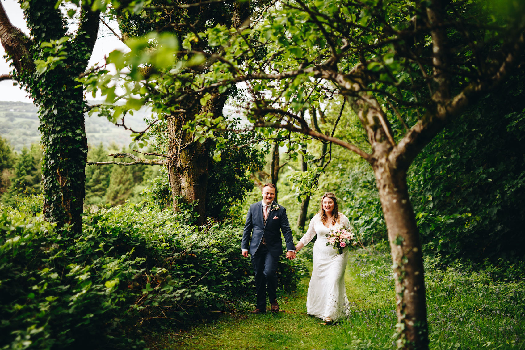 Bride and groom walking towards the camera hand in hand along a path through leafy trees