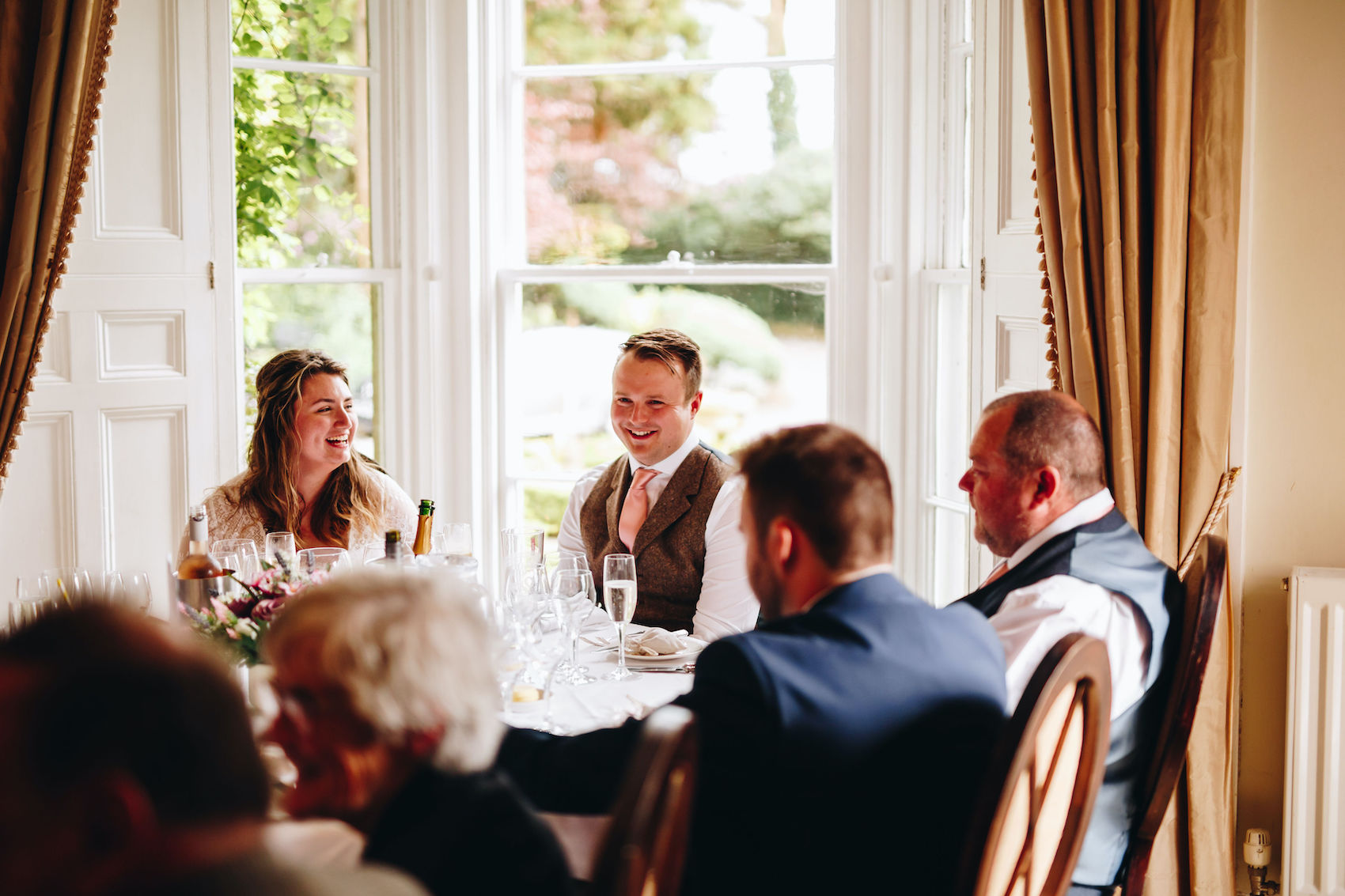 Bride looks on at groom as he laughs at what a guest has said, while seated at their reception