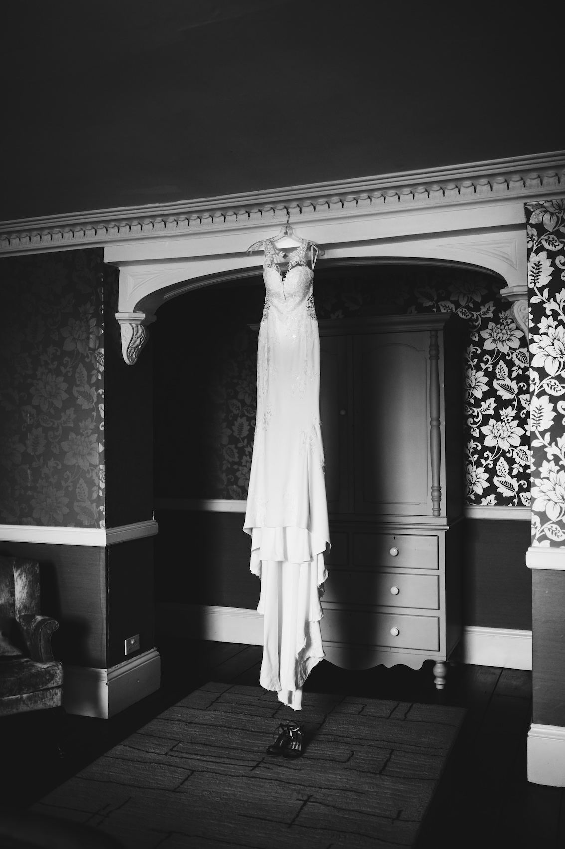 Black and white photo of bride's gown hanging from the ceiling in a beautoifully wallpapered hallway