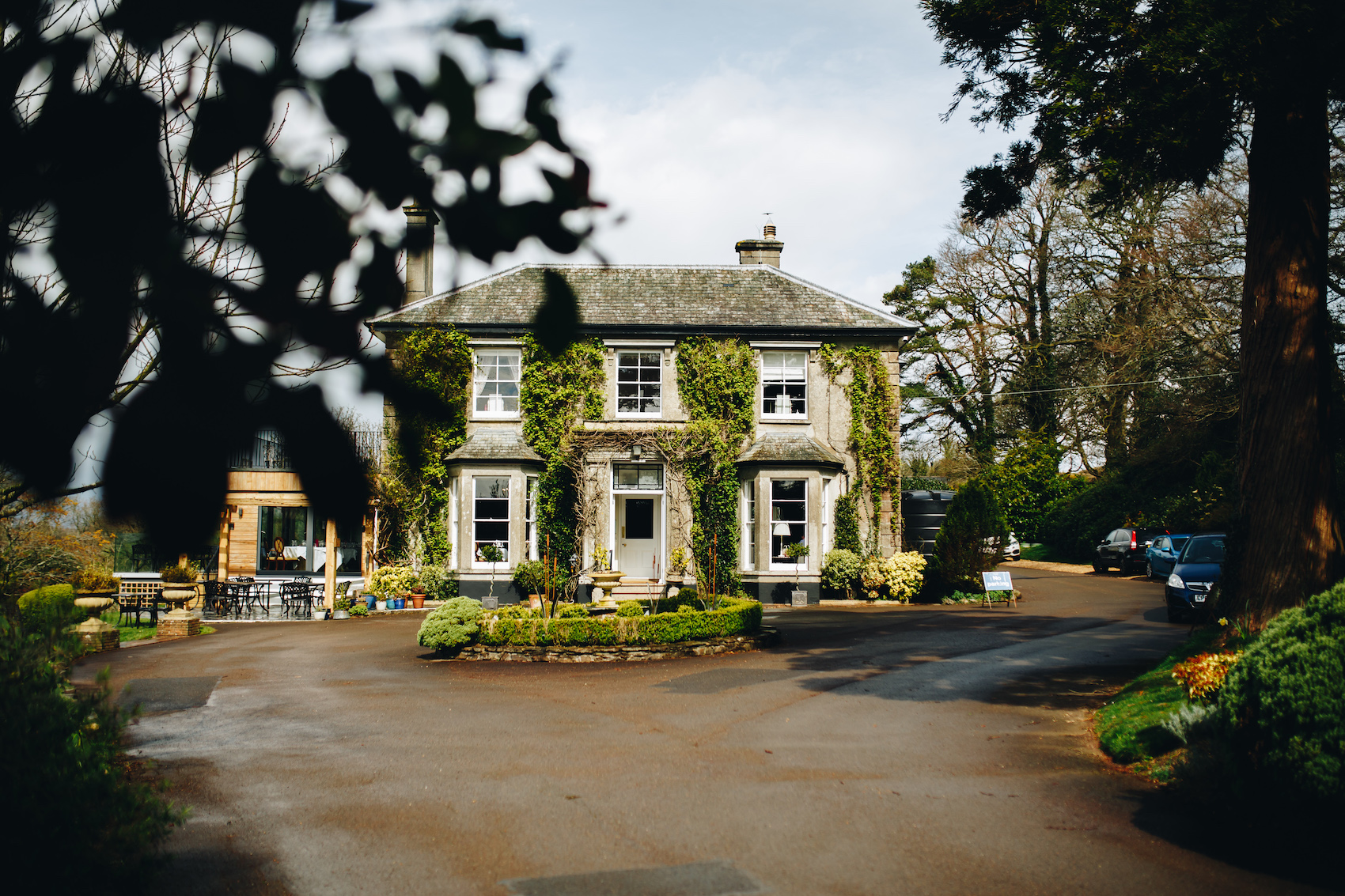 View from the driveway of The Horn of Plenty, through the trees, with its fountain in front. Big building with the front door central, a window on either side and 3 windows upstairs.