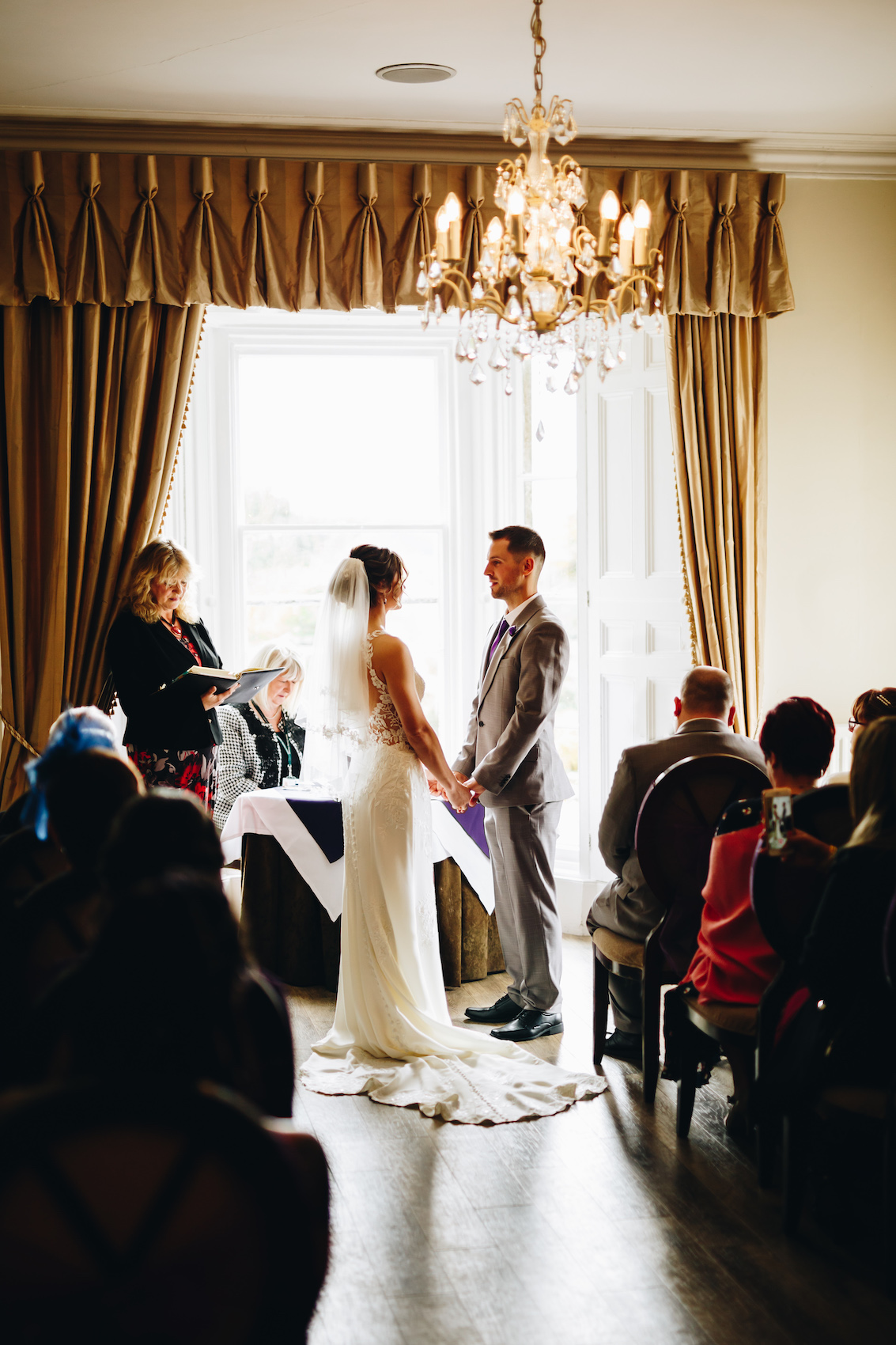 Bride and groom stood hand in hand at the top of the aisle while getting married, by a large curtained window