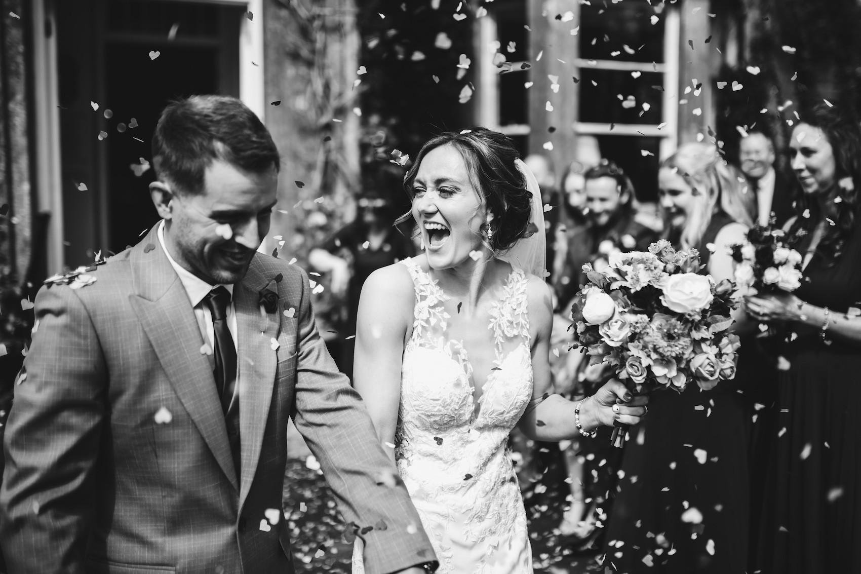 Black and white photo of the bride and groom smiling as they walk out of the building into the shower of confetti