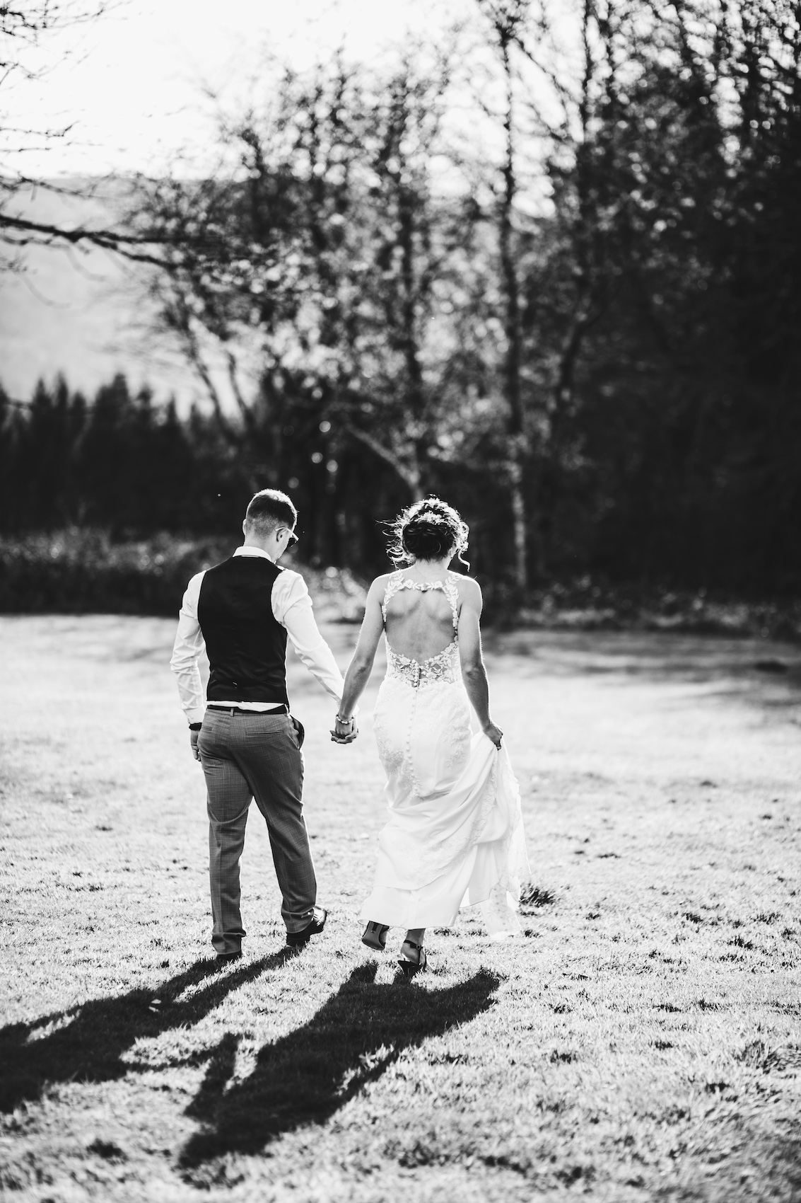Black and white photo of bride and groom walking away through a field towards trees in the evening light