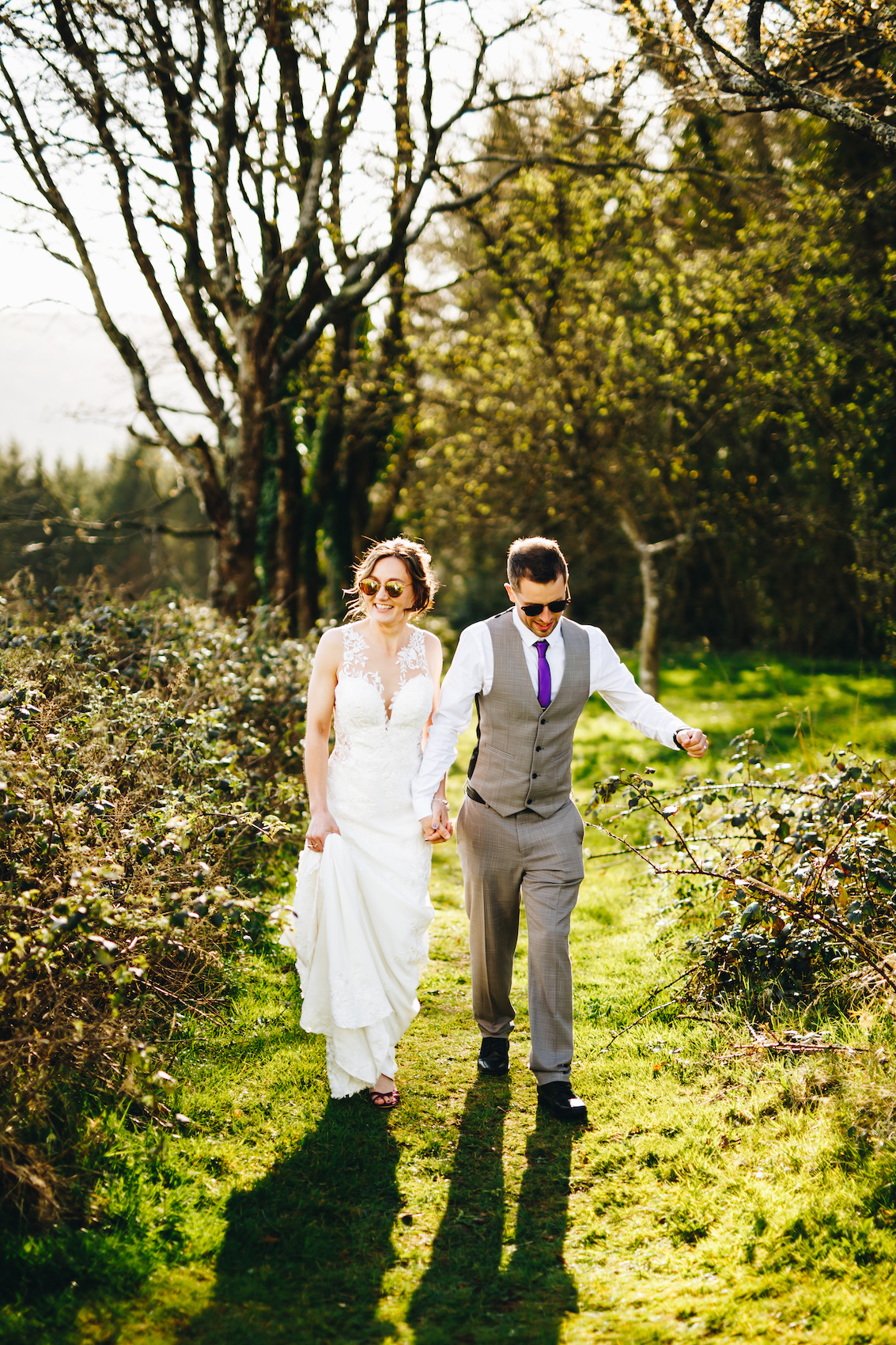 Bride and groom wear sunglasses, walking towards the camera, amongst trees at sunset
