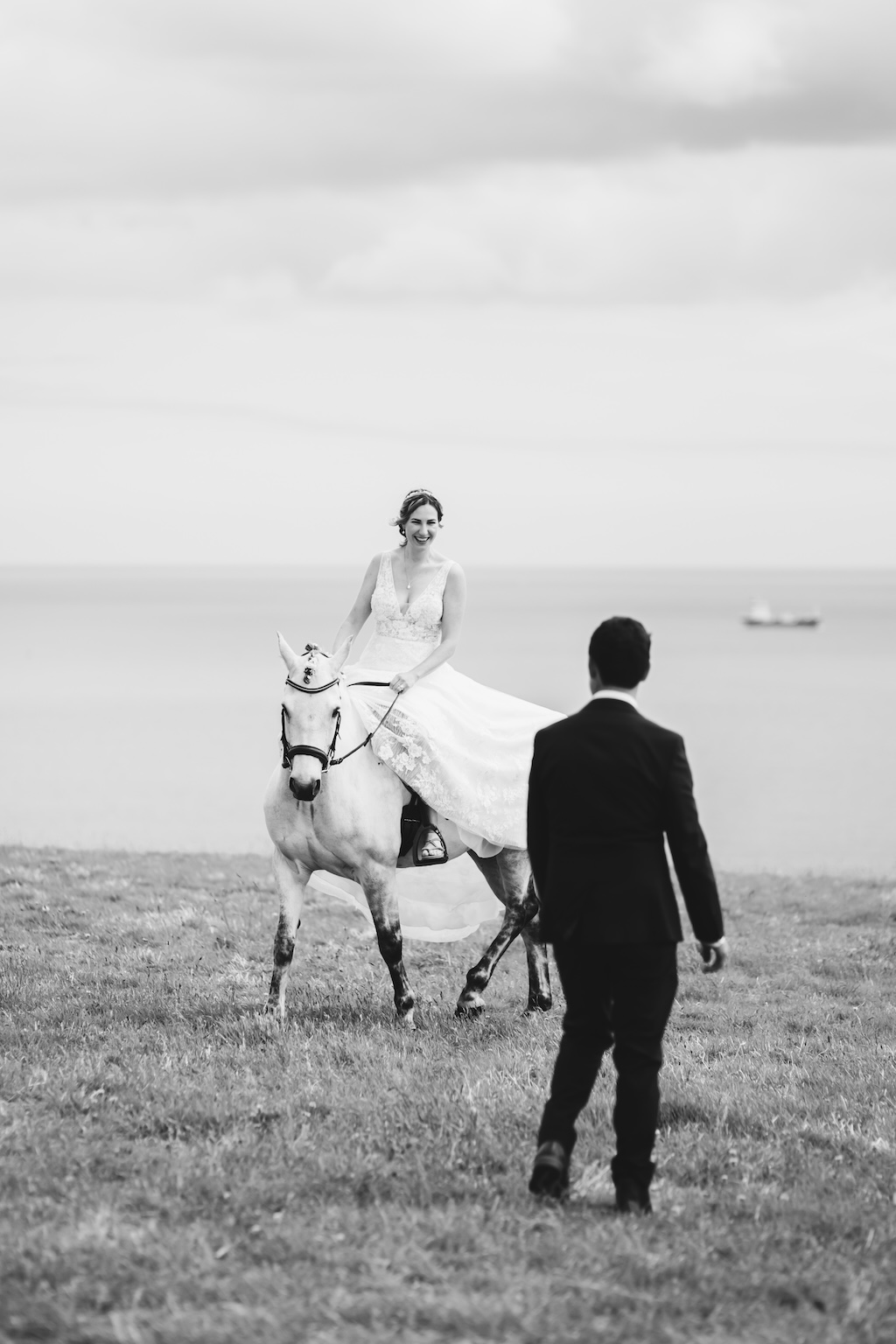 Black and white photo of groom with his back to the camera, looking at his new bride who is riding her white horse.