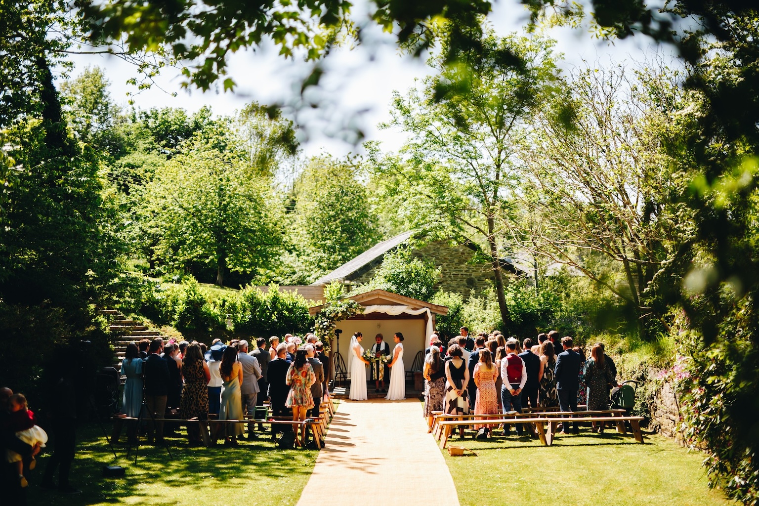 Photo of wooden structure where two brides are getting married, with their friends and family stood by long wooden benches, surrounded by trees and greenery