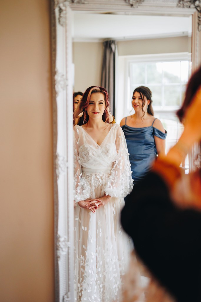 Bride looking happily at her reflection in a full length mirror as her veil gets attached